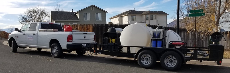 White pickup truck towing a trailer with large water tanks and cleaning equipment, parked on a residential street.