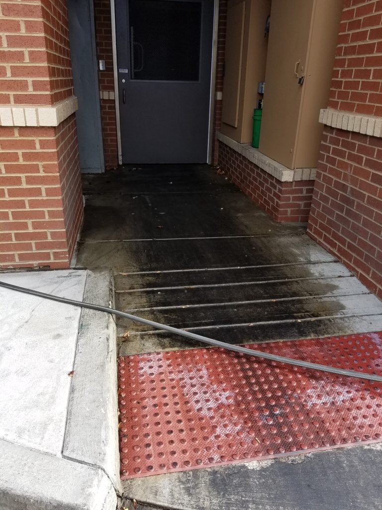A black metal pipe obstructs the entrance ramp to a building with red brick walls and a gray door.