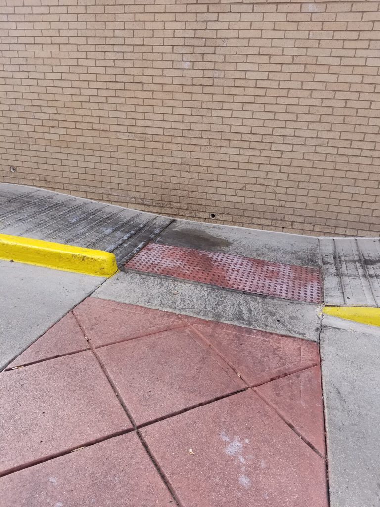 A sidewalk ramp with a textured red surface leads to a brick wall, bordered by yellow-painted curbs.