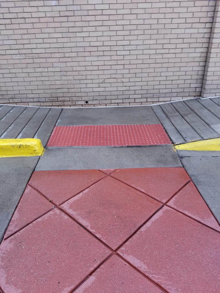 A sidewalk ramp with a red tactile paving panel leads to a brick wall, bordered by yellow painted curbs.